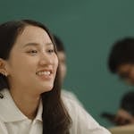 Asian woman smiling while studying at a desk in a classroom setting.