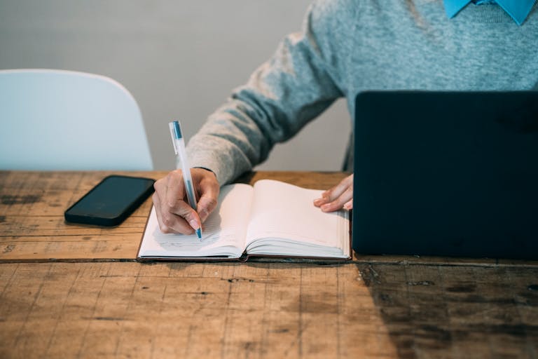 Author writing in notebook in front of laptop and a smartphone at a wooden table with gray background