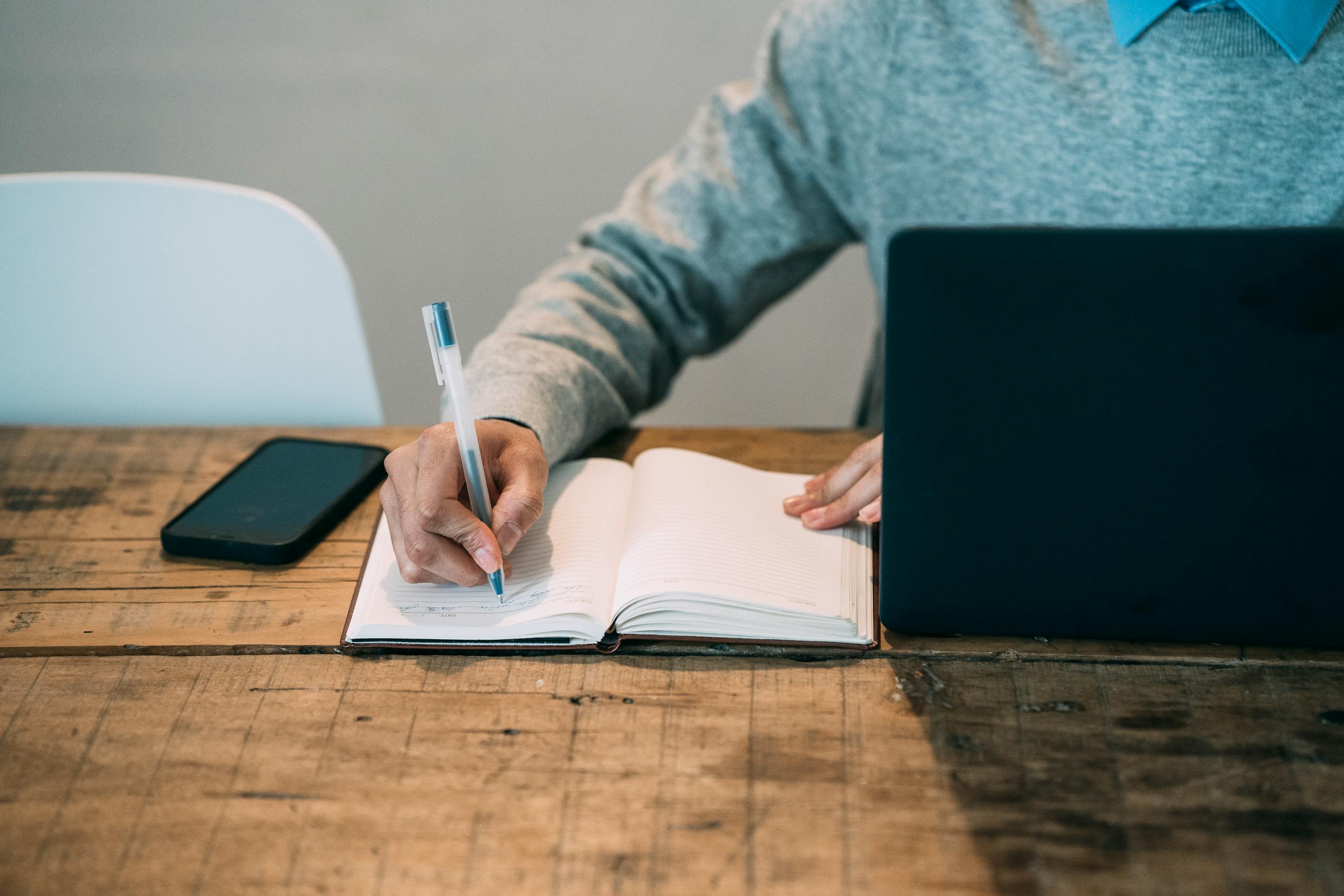 Author writing in notebook in front of laptop and a smartphone at a wooden table with gray background