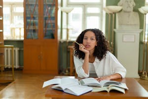 Focused woman with curly hair studying at a library desk filled with open books.