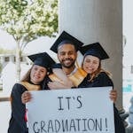 Group of diverse graduates celebrating outdoors with a graduation poster, symbolizing achievement and joy.