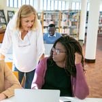 Multiracial students using laptop for studies and doing task while consulting with teacher in university library