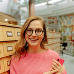 Young woman smiling in a library with a smartphone and eyeglasses, wearing a pink sweater.